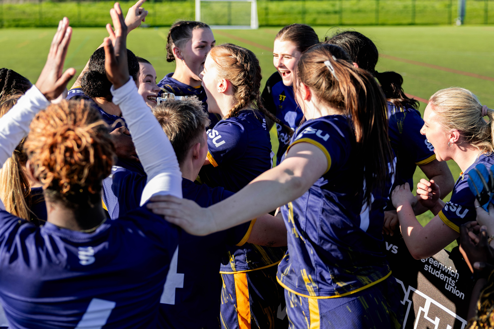womens football team huddled celebrating a win, everyone is cheering and some have hands in the air clapping